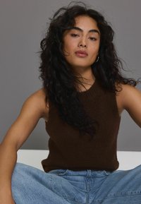 Brauner, ärmelloser Strickpullover mit geripptem Halsausschnitt, kombiniert mit hellblauen, locker sitzenden Jeans. Strukturierter Stoff sichtbar.