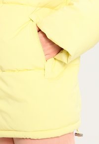 Chaqueta acolchada de color amarillo claro con una textura suave, que cuenta con un bolsillo con cremallera y puños elásticos para mayor comodidad.