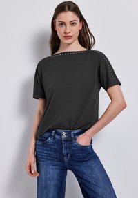 T-shirt à manches courtes gris foncé avec un large décolleté et des clous argentés le long de la couture de l'épaule, assorti à un jean en denim bleu.