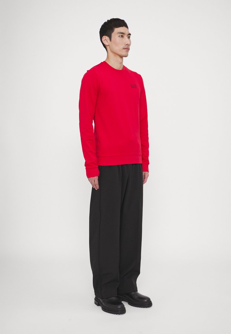 Felpa rossa a maniche lunghe con polsini a coste e scollatura rotonda, con un piccolo logo nero. Abbinata a pantaloni neri larghi e scarpe nere.