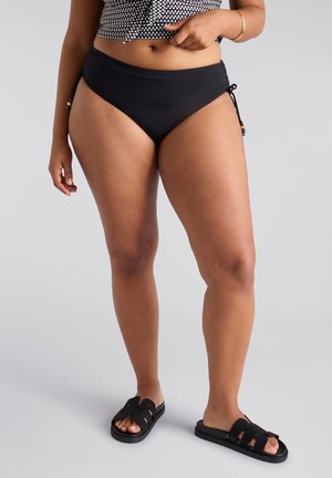 Parte inferior de bikini negra con lazo ajustable a los lados y adornos decorativos en oro. Tejido suave con cobertura moderada, combinada con sandalias deslizantes negras.