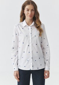 Witte overhemd met lange mouwen en blauwe bloemenbroderie, knoopsluiting aan de voorkant, kraag en zachte stof. Draagt men bij donkere spijkerbroek.