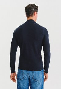 Granatowa, długorękawowa koszulka polo o dopasowanym kroju, z prążkowanymi ściągaczami i gładką fakturą. Noszona z jasnoniebieskimi dżinsami.