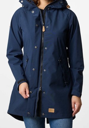 Person iført en mørkeblå hætte-regnjakke med frontlynlås og trykknapper, kombineret med blå jeans, på en ensfarvet baggrund.