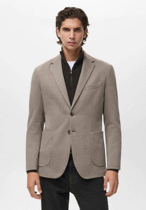 Blazer de mezclilla de lana beige con un ajuste entallado, solapas de muesca, dos bolsillos frontales y textura contrastante. Llevado sobre un suéter oscuro con cremallera.