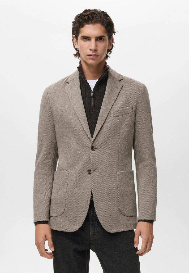 Blazer in misto lana beige con una vestibilità su misura, rever a punta, due tasche frontali e texture a contrasto. Indossato su un maglione scuro con zip.