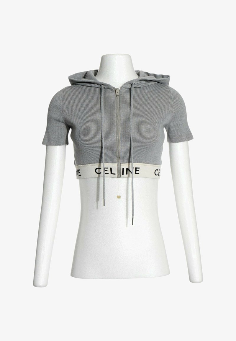 Sweat à capuche court gris manches courtes avec fermeture éclair devant, cordons et bande élastique blanche à l'ourlet avec texte noir "CELINE" sur un mannequin blanc.