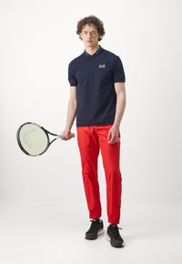 Navy polo shirt with collar and logo, red tracksuit pants, black sneakers. Model holding a tennis racket, standing on a white background.