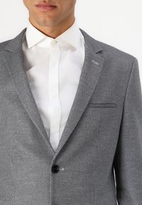 Grå mønstrete blazer med notched lapel, enkel knappelukking og teksturert stoff, båret over en hvit skjorte med krage.