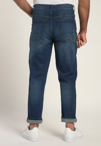 Jeans da uomo blu scuro con vestibilità affusolata, cuciture visibili e due tasche posteriori. Polsini arrotolati sopra sneakers bianche.