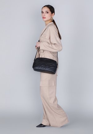 Borsa a tracolla in pelle nera con una texture liscia, design trapuntato e dettagli borchiati, indossata crossbody con una lunga tracolla.
