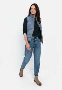 Gepolsterte graue Weste mit hohem Kragen über einem marineblauen Sweatshirt, kombiniert mit Jeans in mittlerem Waschton und braunen Stiefeletten, die eine lockere Passform aufweisen.