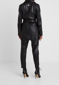 Svart läderjacka med bälte i midjan, med en sydd etikett på baksidan, kombinerad med åtsittande svarta lädershorts och klackboots.