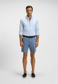 Light blue striped dress shirt with buttoned collar, paired with solid blue shorts and dark slip-on shoes. Man stands against a neutral background.