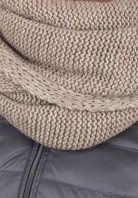 Écharpe beige en maille à infinity avec un motif texturé et épais, superposée sur une doudoune gris foncé avec un détail de fermeture éclair.