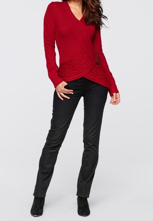 Donna che indossa un maglione rosso a fascia in maglia a trecce con dettaglio fibbia, pantaloni neri aderenti e stivaletti neri su uno sfondo semplice.