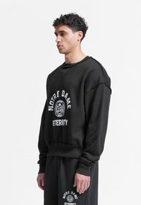 Νέος άνδρας με μαύρο φούτερ της Αδελφότητας Notre Dame και αντίστοιχο παντελόνι, στέκεται σε προφίλ μπροστά σε λευκό φόντο.