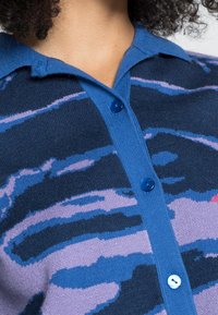 Pletená polokošile v tmavě modré barvě s fialovým abstraktním vzorem; má modrý límec a légu na knoflíky s modrými knoflíky.