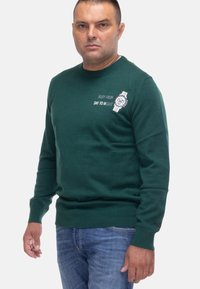 Maglione girocollo verde con testo "IMPEGNATO DAL MATTINO ALLA SERA" e grafica di un orologio. Texture morbida, polsini e orlo a coste. Abbinato a jeans azzurri chiari.