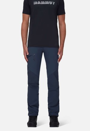 Mand iført mørkeblå outdoorbukser med lynlåslomme på låret, sorte vandrestøvler og sort T-shirt med "Mammut" logo.