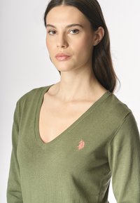 Modello con capelli lunghi che indossa un maglione verde oliva a scollo a V con un piccolo logo ricamato sul petto, su uno sfondo semplice.