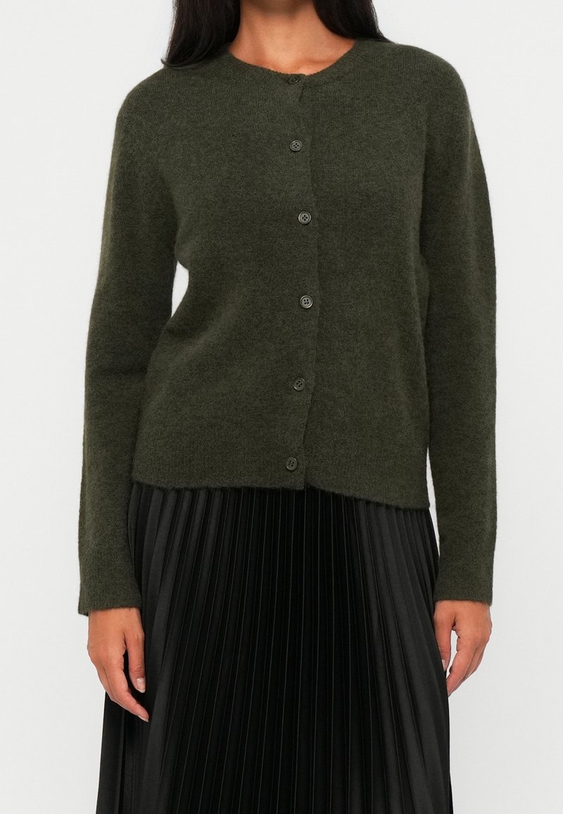 Femme portant un cardigan boutonné vert foncé associé à une jupe plissée noire, visible des épaules jusqu'au milieu de la cuisse.