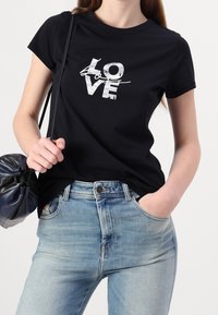 Mujer vistiendo una camiseta negra con el texto blanco "AMOR" y unos jeans azul claro, sosteniendo un bolso de hombro negro y azul marino.