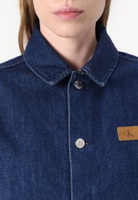 Džinsinis švarkas, pasižymintis tamsiai mėlyna spalva, mygtukų uždarymu, klasikiniais apykaklėmis ir odiniu logotipo ženkliuku krūtinėje.