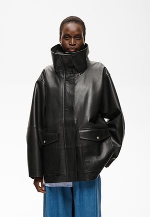 Persoon die een oversized zwarte leren jas draagt met grote voorzakken en een hoge kraag, gecombineerd met losse blauwe broek.