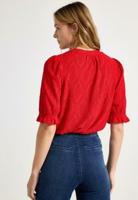 Kvinde vendt mod venstre iført en rød tekstureret bluse med pufærmer og mørkeblå højtaljede jeans mod en ensfarvet baggrund.