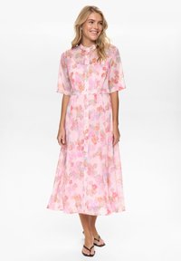 Usmievavá žena v ružových kvetovaných midi šatách na gombíky s lakťovými rukávmi a čiernych sandáloch na bielom pozadí.