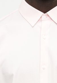 Κοντινό πλάνο ενός ατόμου με σκούρο δέρμα που φοράει ένα ανοιχτό ροζ πουκάμισο με κουμπιά και μυτερό γιακά.