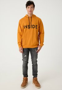 Sudadera con capucha en mostaza con impresión en negro "INSIDE", bolsillo frontal. Combinada con jeans gris desgastados y botas beige con cordones.