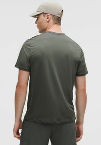 T-shirt de sport à manches courtes, vert foncé, fabriqué en tissu lisse. Le mannequin porte une casquette beige, arborant un design simple et ajusté.