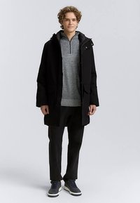 Svart lång kappa med huva, försedd med två framfickor och tryckknappar. Bärs över en grå tröja och matchas med mörka byxor och sneakers.