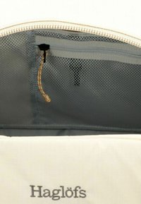 Intérieur d'un sac textile beige avec une poche en maille grise, présentant une fermeture éclair, un cordon de tirage jaune et rouge, et le logo Haglöfs sur le tissu extérieur.