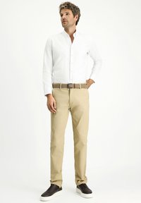 Camicia bianca a maniche lunghe con bottoni, chinos beige, cintura intrecciata marrone e scarpe slip-on scure con suole bianche, in piedi contro uno sfondo neutro.