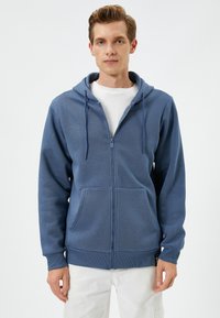 Sudadera azul con cremallera, hecha de una tela suave, que cuenta con una capucha ajustable, bolsillos frontales y puños de canalé. Textura suave con un patrón sutil.