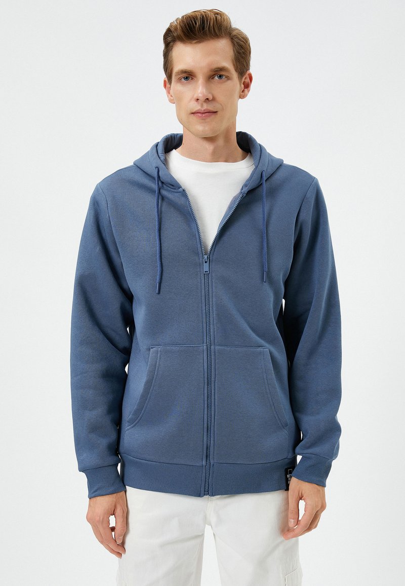 Sudadera azul con cremallera, hecha de una tela suave, que cuenta con una capucha ajustable, bolsillos frontales y puños de canalé. Textura suave con un patrón sutil.