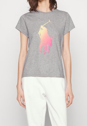 Donna che indossa una t-shirt grigia con un logo di un giocatore di polo sfumato rosa e giallo sul petto, abbinata a pantaloni bianchi.