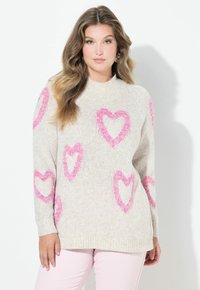 Junge Frau mit langen welligen Haaren, die einen cremefarbenen Pullover mit pinken Herzmustern und hellrosa Hosen trägt und vor einem weißen Hintergrund steht.