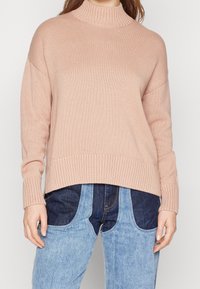 Eine Frau trägt einen lockeren, gerippten, pinkfarbenen Pullover, kombiniert mit zweifarbigen blauen Jeans aus Denim mit einem auffälligen Patch-Design auf der Vorderseite.