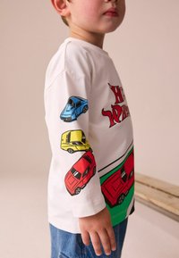Biele tričko s dlhým rukávom s farebnými grafickými motívmi áut v modrej, červenej a žltej farbe, okrúhlym výstrihom a jemnou bavlnenou textúrou.