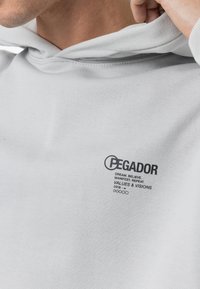Lys grå hoodie med en kengurulomme, ribbede manchetter og et logo med tekst på brystet, lavet af blød bomuldsmateriale.