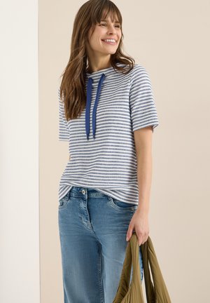 Femme souriante portant un haut à rayures bleues à manches courtes et un jean, tenant un sac fourre-tout vert olive sur un fond neutre.