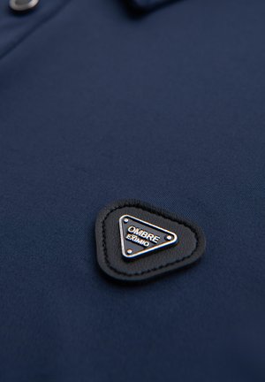 Granatowa koszulka polo o gładkiej fakturze, z trójkątną czarną skórzaną łatą z białym napisem "OMBRE" oraz srebrnymi akcentami.