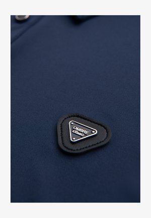 Granatowa koszulka polo o gładkiej fakturze, z trójkątną czarną skórzaną łatą z białym napisem "OMBRE" oraz srebrnymi akcentami.