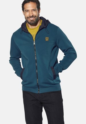Teal zip-up hoodie with a navy-lined hood, front pockets, and a small logo patch. Paired with a yellow shirt and dark jeans.