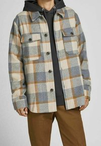 Giacca a quadri con motivi beige, blu e marroni, dotata di due tasche frontali, chiusura con bottoni e una vestibilità leggermente oversize sopra un felpa grigia.