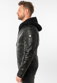 Giacca di pelle nera con cappuccio testurizzato nero, chiusura con zip sul davanti e una patch con logo sulla spalla. Finitura liscia con design aderente.